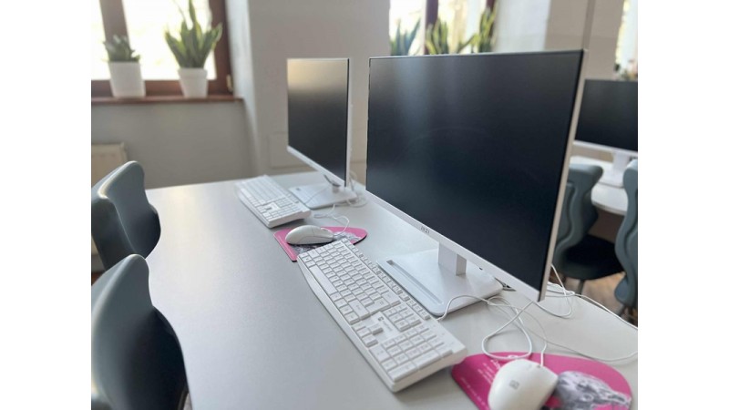 Rząd stanowisk komputerowych w jasnej sali: monitory, klawiatury i myszy ustawione na biurkach.